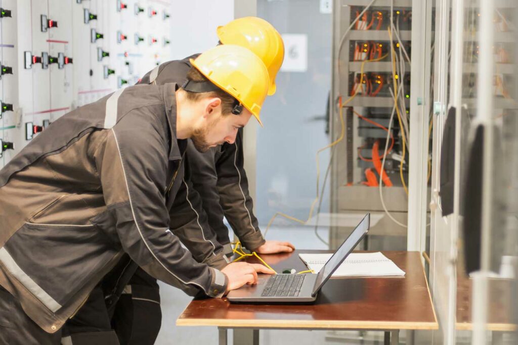Two field technicians in hard hats using a laptop for equipment diagnostics in an industrial environment