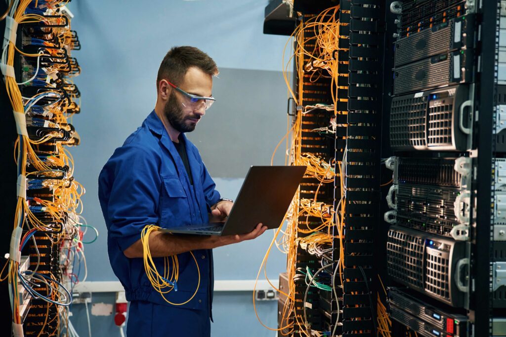 Technician wearing protective glasses and blue overalls working on a laptop in a server room, surrounded by racks of network servers and cables.